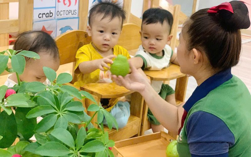 Cô trao đồ vật cho bé nhà trẻ quanh bàn ăn — mỗi lần trao là một lần giao tiếp